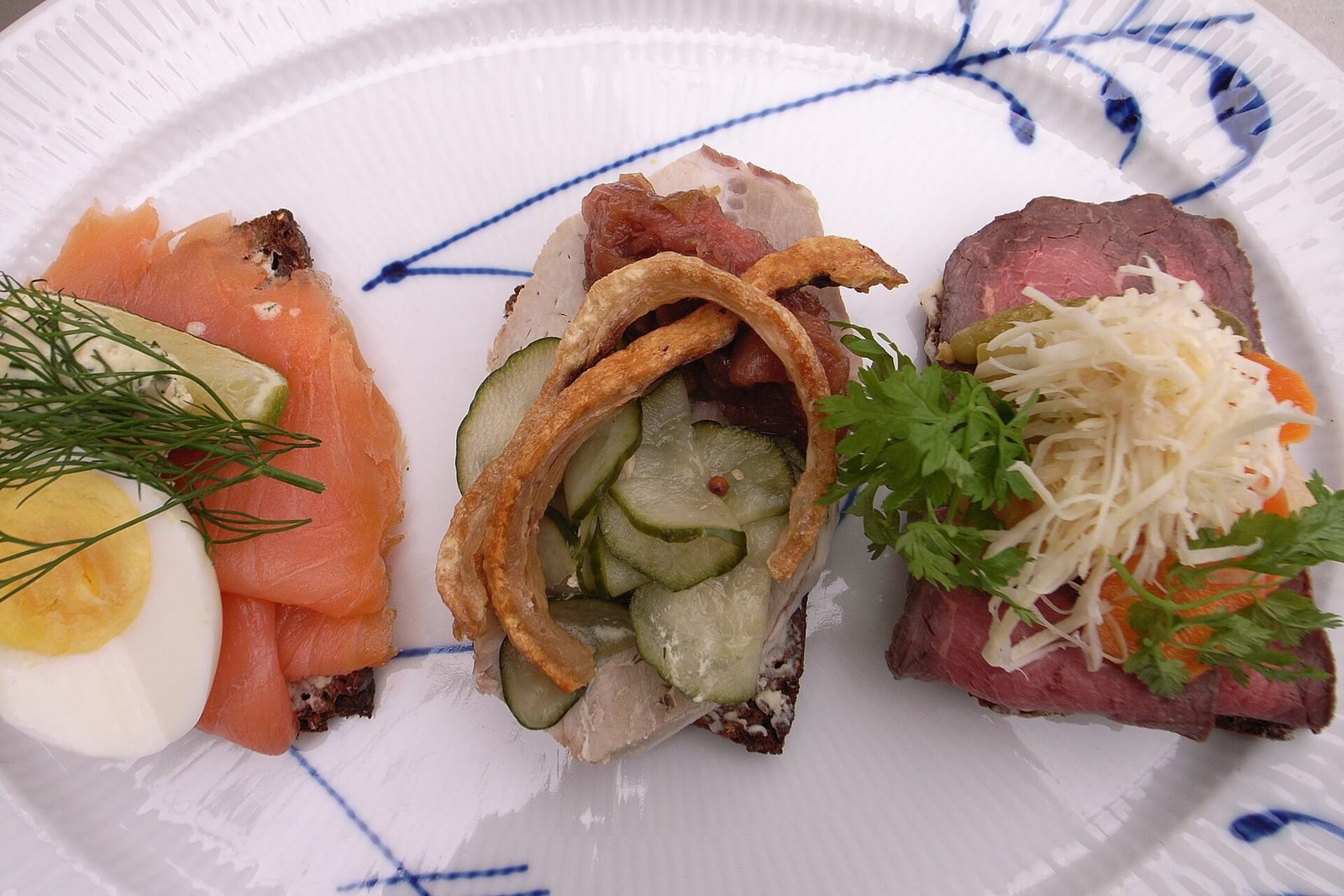 Tre stykker sm&oslash;rrebr&oslash;d med laks og &aelig;g, sild og roastbeef p&aring; en bl&aring;m&oslash;nstret tallerken